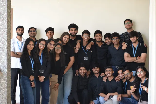 A large group of students in black, many wearing university lanyards, posing against a white wall