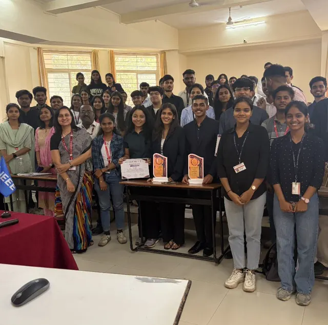 A large group of students in a classroom with an IEEE flag, a certificate and two trophies on the table